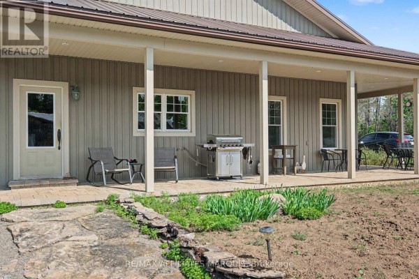 View of the house's wrap-around porch, showing a grill, chairs, and a stone pathway leading to a door. - 14222 County Rd 15 Road E, Merrickville Wolford