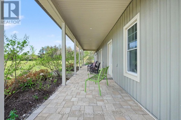 Long shot of the covered wrap-around porch with light grey siding and stone paver flooring, overlooking a green field. - 14222 County Rd 15 Road E, Merrickville Wolford