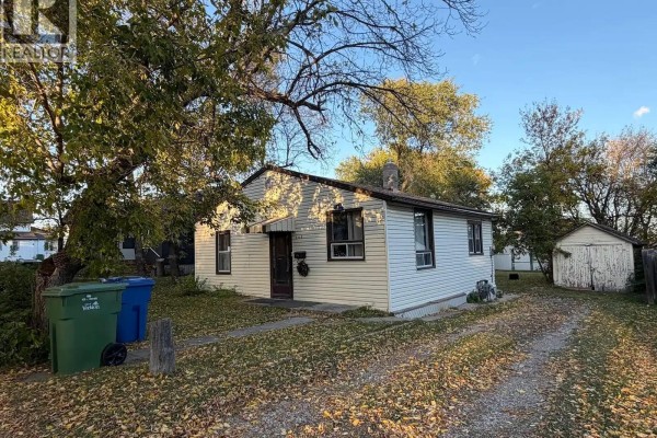 Front exterior of a small home with a driveway, trees, and garbage bins. - 143 Sixth Avenue N, Yorkton