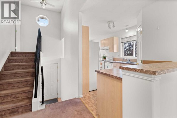 Interior view showing a staircase with brown carpet, a white door, and a glimpse of the kitchen. - 144, 55 Fairways Drive Nw, Airdrie