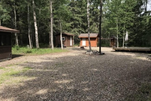 Open lot with gravel surface, two cabins, and a deck, surrounded by trees. - 144 Ahlstrom Drive, Turtle Lake