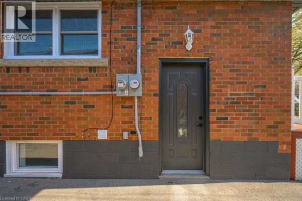 Exterior brick wall with a grey door, electrical meters, and a basement window. - 144 East 44th Street Unit# Lower Unit, Hamilton