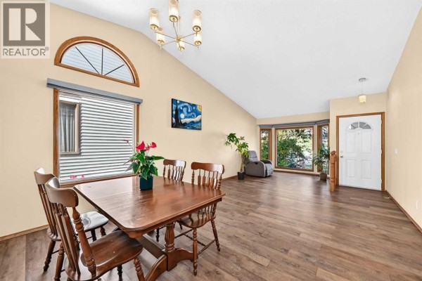 Dining and living area with a wooden table, vaulted ceiling, and bay windows. - 144 Hawkdale Close Nw, Calgary