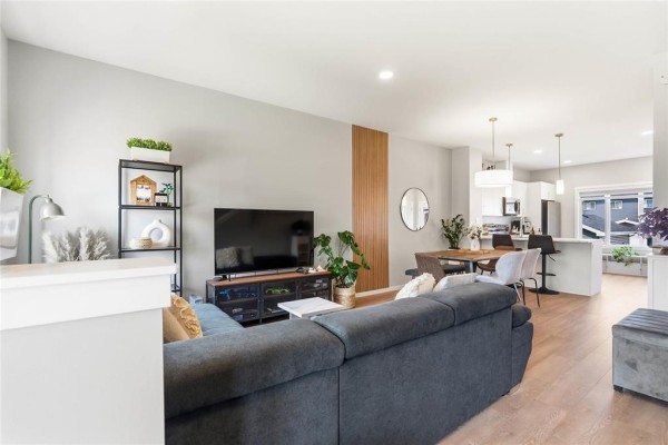 Living room with a gray sectional sofa, television, shelves, and an open-plan view to the dining area and kitchen. - 146 Emberwell Street, Winnipeg