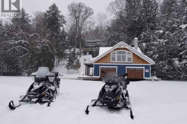 Two snowmobiles parked on a snow-covered lake in front of a blue boathouse. - 146 Maplehurst Road, Seguin