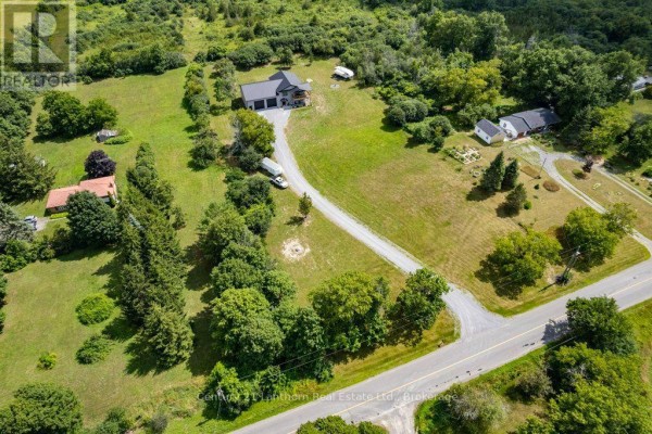 Aerial view of a property with a grey house, garage, driveway, and surrounding green landscape. - 1467 County Road 19, Prince Edward County