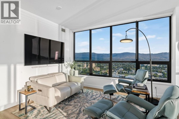 Living room with large windows, two reclining chairs, a sofa, and a view of the city and mountains. - 1488 Bertram Street Unit# 3202, Kelowna