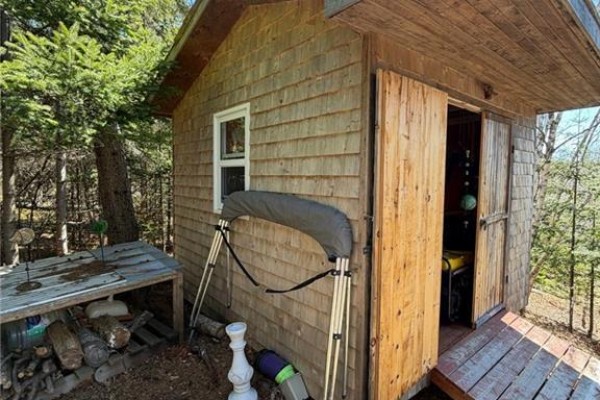 Side view of a wooden shed with large double doors, a small window, and outdoor storage including logs and a boat canopy. - 149 Limekiln Road, Letang