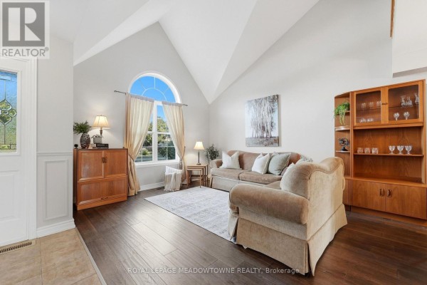 Living room with vaulted ceiling, large arched window, beige sofa, armchair, and dark hardwood floors. - 15 Costigan Court, Halton Hills