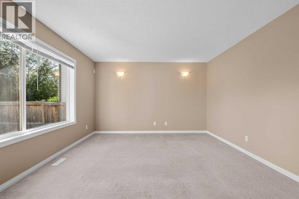 Empty room with light brown walls, beige carpet, and a large window showing greenery outside. - 15 Cranfield Manor Se, Calgary