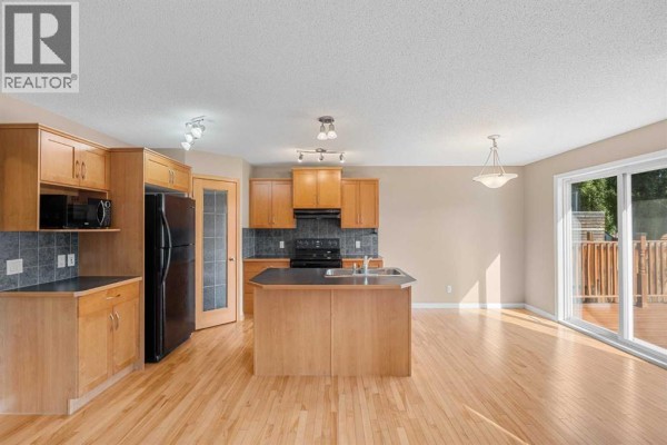 Kitchen with light wood cabinets, black appliances, a central island, and adjacent dining area with a sliding glass door. - 15 Cranfield Manor Se, Calgary