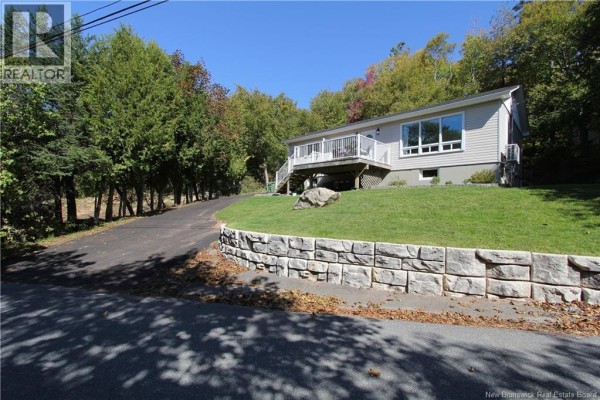 View from the driveway of a white house with a large lawn, retaining wall, and wooded surroundings. - 15 Hazen Avenue, Rothesay