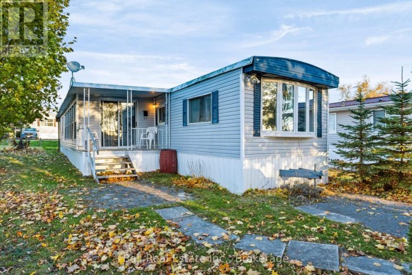 A light blue mobile home with a covered porch and bay window, surrounded by autumn leaves. - 15 Mcgregor Central Drive, Otonabee South Monaghan
