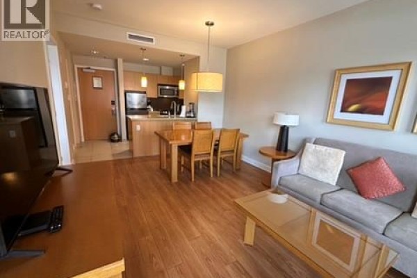 Living and dining area with grey sofa, wooden table, and television on wooden floors. - 15 Park Place Unit# 209, Osoyoos