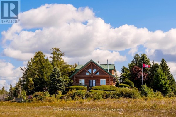 A log cabin with a large arched window on the front facade, seen from a grassy field with trees and a Canadian flag. - 150 Shoreline Avenue, South Bruce Peninsula