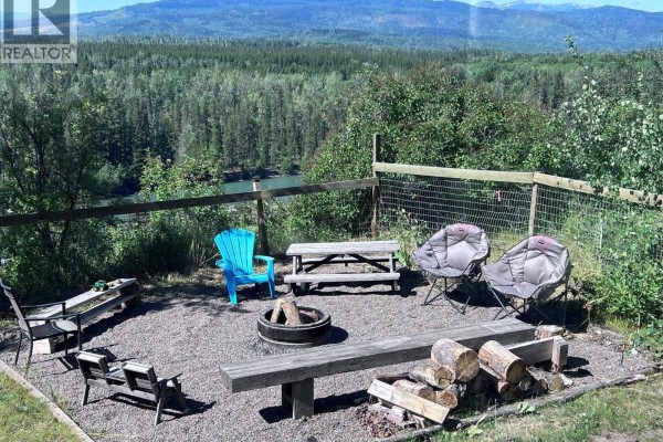 Outdoor fire pit area with gravel ground, wooden benches, chairs, a picnic table, and firewood pile. - 1500 2nd St. (aldermere Ridge), Telkwa