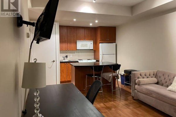 Living area with a grey sofa, desk with chair, mounted TV, and kitchen with wood cabinets. - 1501 - 85 Bloor Street E, Toronto