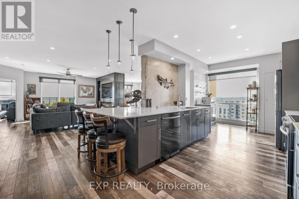 Island kitchen with grey cabinets, concrete accent wall, and seating for three bar stools. - 1502 - 65 Ellen Street, Barrie