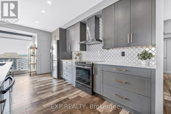 Modern kitchen with grey flat-panel cabinets, stainless steel appliances, and white patterned backsplash. - 1502 - 65 Ellen Street, Barrie