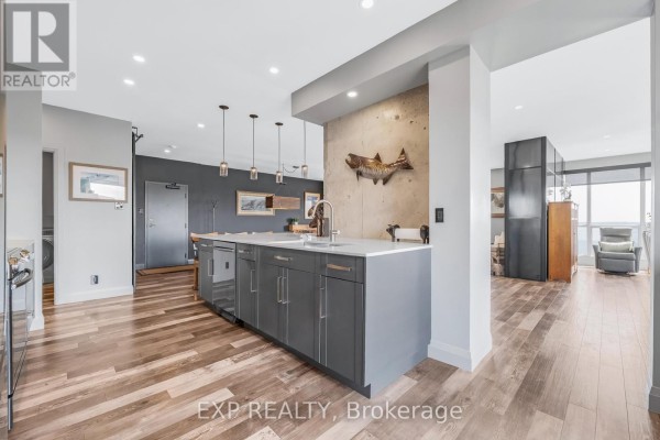 Kitchen island with grey cabinets and a sink, open to a living area with large windows. - 1502 - 65 Ellen Street, Barrie