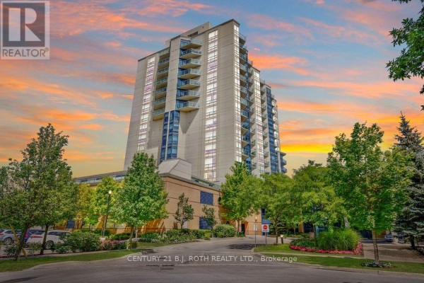 Exterior of a beige apartment building with glass balconies at sunset with trees. - 1505 - 37 Ellen Street, Barrie