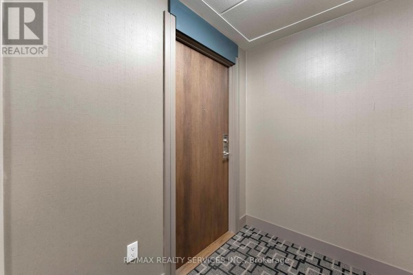 Entryway with a wooden door, a black doormat, and a view into a kitchen with stainless steel appliances. - 1506 - 3975 Grand Park Drive, Mississauga