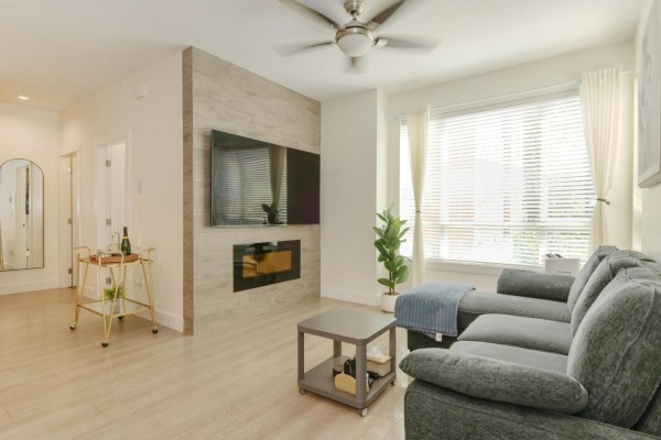 Living room featuring a large grey sectional sofa, gas fireplace, mounted television, and a window with white curtains. - 151 16433 19 Avenue, Surrey