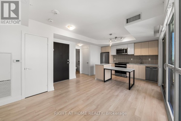 Living area and kitchen with dark front door, stainless steel appliances, and island. - 1511 - 1461 Lawrence Avenue W, Toronto