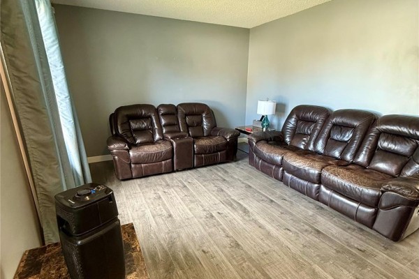 Living room with two reclining brown leather sofas, grey walls, and light wood-look vinyl flooring. A teal curtain hangs on the left. - 1511 Grandview Street W, Moose Jaw
