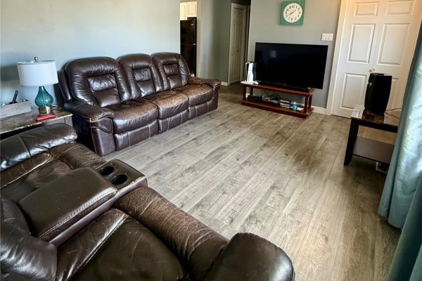 Living room with a sectional brown leather sofa, television on a media console, and a clock on the wall. Light wood-look vinyl flooring. - 1511 Grandview Street W, Moose Jaw