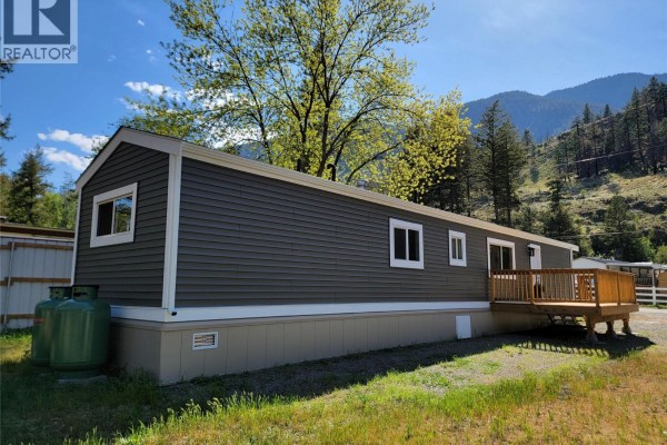 Right side view of a grey mobile home with white trim, propane tanks, and a mountainous, wooded background. - 1514 Highway 3a Unit# 3, Keremeos