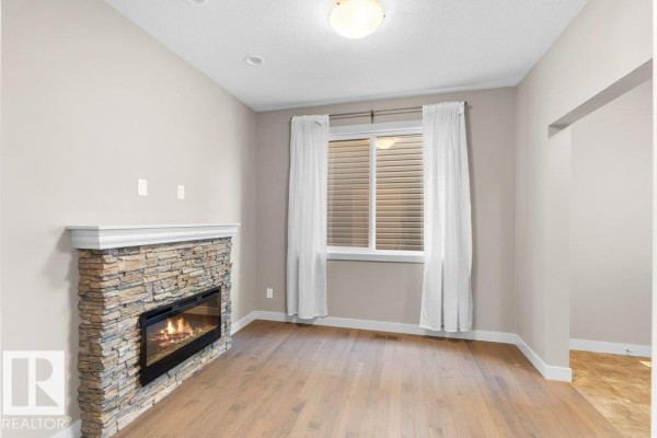 Living room with light wood floors, a stone gas fireplace, and a large window with white curtains. - 1523 168 St Sw, Edmonton