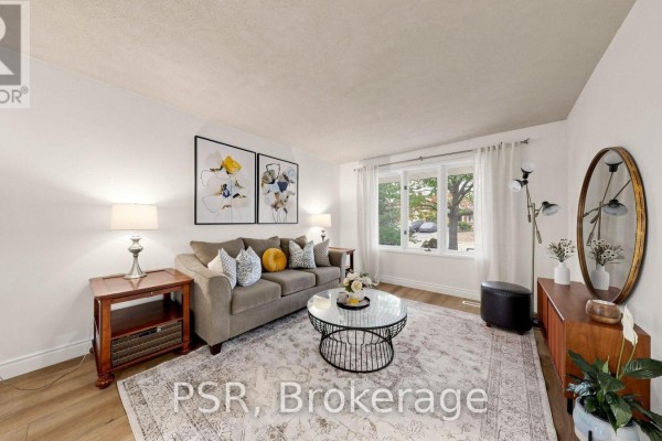 Living room with a sofa, coffee table, round mirror, and large bay window overlooking trees. - 1528 Wildlark Drive, Peterborough
