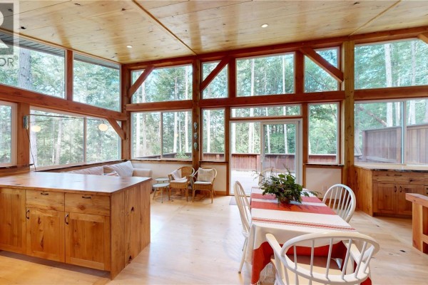 A dining table with white chairs is set up in a sunroom with wood beams and forest views. - 221 Sockeye Dr, Mudge Island