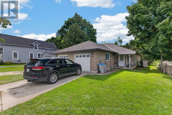 Side exterior view of a duplex with two garage doors, tan siding, a green lawn, and trees. - 154&156 Princess Street E, Central Huron