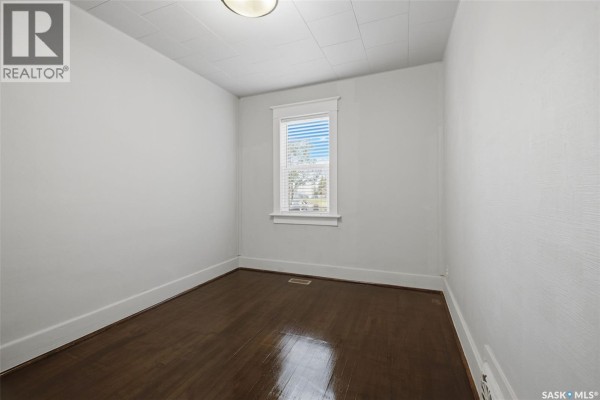 Empty bedroom with dark hardwood floors, white walls, and a window with blinds. - 1545 Princess Street, Regina