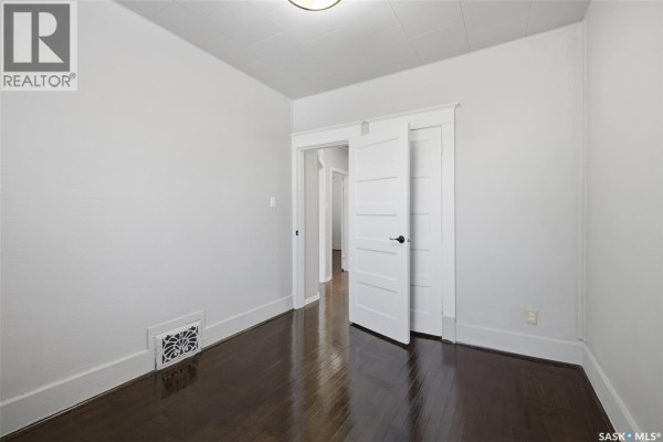 Empty bedroom with dark hardwood floors, white walls, open closet doors, and a window with blinds. - 1545 Princess Street, Regina