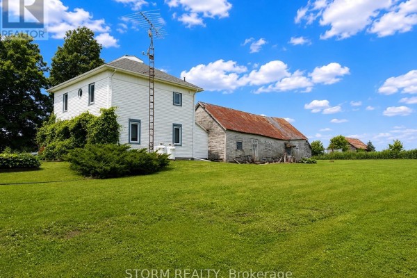 White house with a large barn next to it on a grassy field with trees in the background. - 15477 Raymond Road, South Stormont