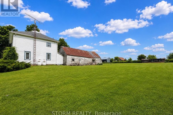 White house and barn in a wide grassy field with trees and blue sky with clouds. - 15477 Raymond Road, South Stormont