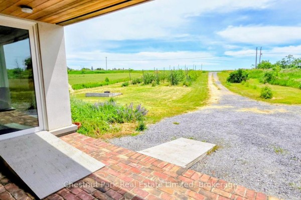 View from a covered porch with brick walkway looking out onto a gravel driveway and fields. - 1558 Gilead Road, Prince Edward County