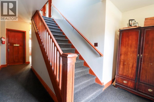 Interior staircase with gray carpet, wooden banisters, and red trim. - 156 Nanaimo Avenue E, Penticton