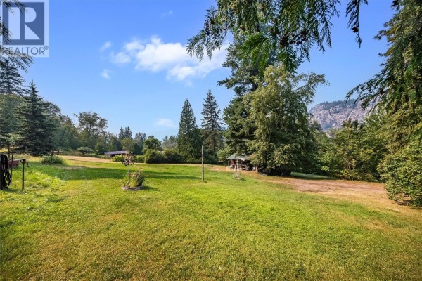 A grassy yard with trees, a small covered structure, and distant mountains under a blue, cloudy sky. - 1562 Arrow Lakes Drive, Castlegar