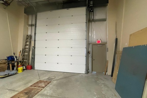 Inside a large commercial bay showing a white roll-up door, a smaller exit door, and stored items. - 15634 - 112 Avenue Nw, Edmonton