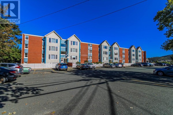 Exterior view of a brick and light blue apartment building with parked cars in the foreground. - 157 Forest Road Unit#65, St  John S