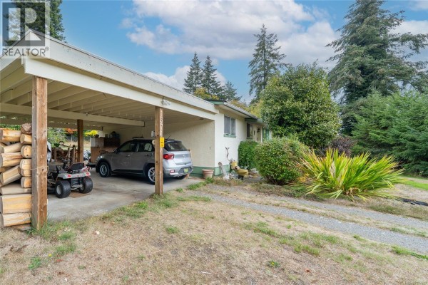Side view of the house with a carport, parked car, lawnmower, and piled firewood. - 1571 Tatlo Rd W, Crofton