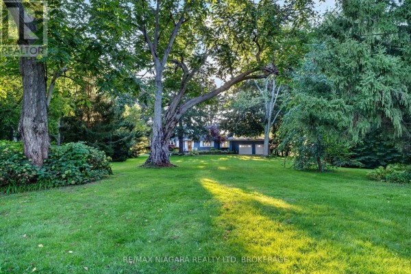 Expansive green lawn with mature trees in the foreground and a long ranch-style home with garage in the distance. - 1577 Gregory Road, St  Catharines