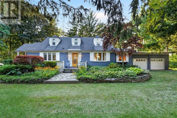 Front facade of a blue ranch home with dormer windows, white railings, a stone walkway, and lush garden beds. - 1577 Gregory Road, St  Catharines