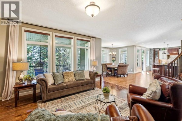 Living room with a brown sofa, leather armchairs, large windows overlooking trees, and hardwood floors. - 158 Hawk's Landing Drive, Rural Foothills County