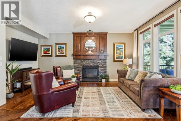 Living room with a stone fireplace, dark wood mantel, brown leather chairs, a sofa, and hardwood floors. - 158 Hawk's Landing Drive, Rural Foothills County