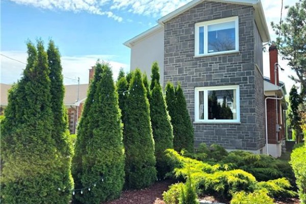 Side exterior of a two-story house with grey stone siding, manicured hedges, and white-trimmed windows. - 16 Forest Hill Road, St  Catharines